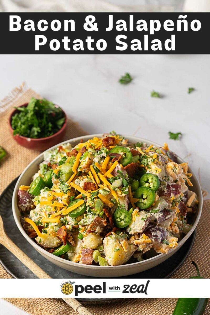 A bowl of jalapeno popper potato salad topped with bacon, jalapeños, shredded cheese, and green onions sits on a white table next to a bowl of chopped herbs.