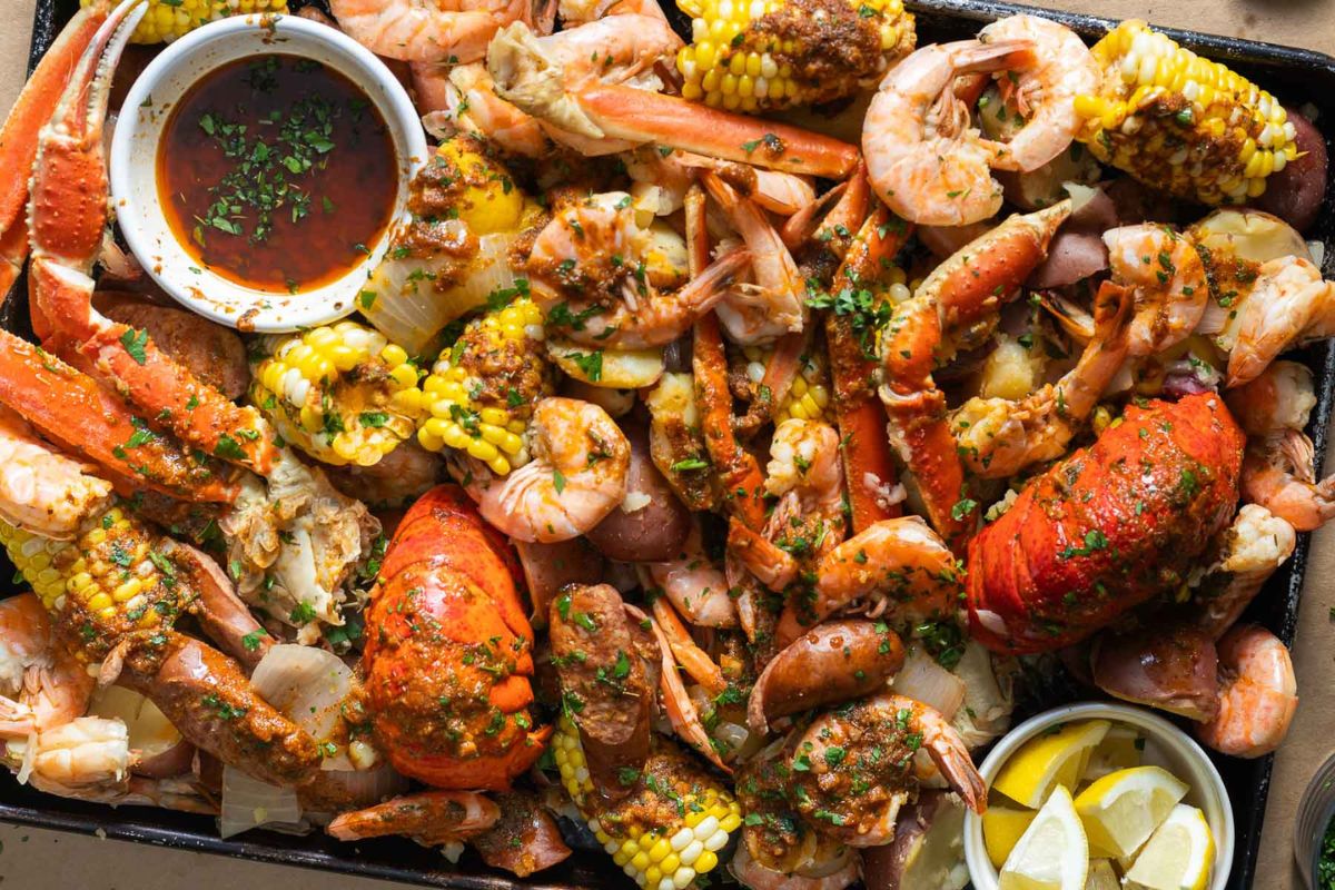 A large tray filled with crab legs, shrimp, lobster, corn on the cob, sausage, potatoes, and lemon wedges brings classic Cajun cuisine to life. Garnished with chopped herbs and served with a bowl of dipping sauce for true Southern flavor.
