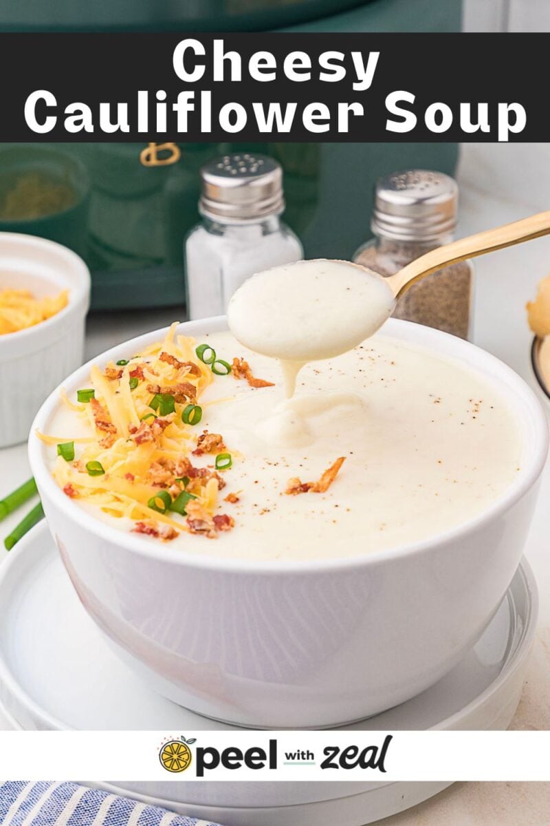 A bowl of cheesy cauliflower soup topped with shredded cheese, green onions, and bacon bits, with a spoon lifting some soup.