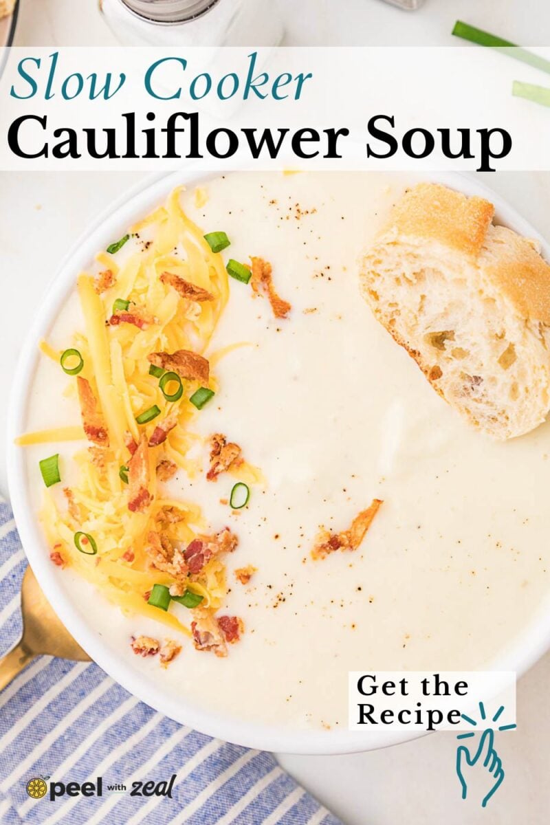 A bowl of creamy cauliflower soup topped with shredded cheese, bacon bits, and green onions, served with a slice of bread.