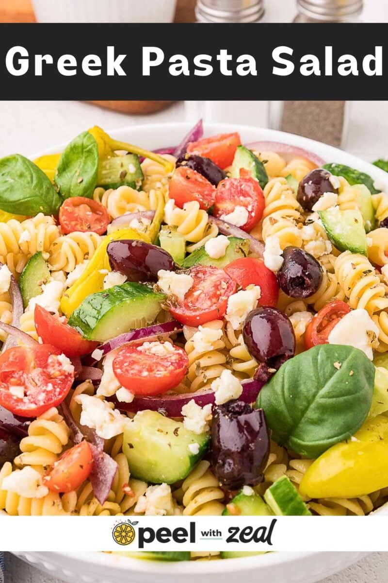 A bowl of Greek pasta salad with rotini, cherry tomatoes, cucumbers, Kalamata olives, red onion, feta cheese, and fresh basil leaves.