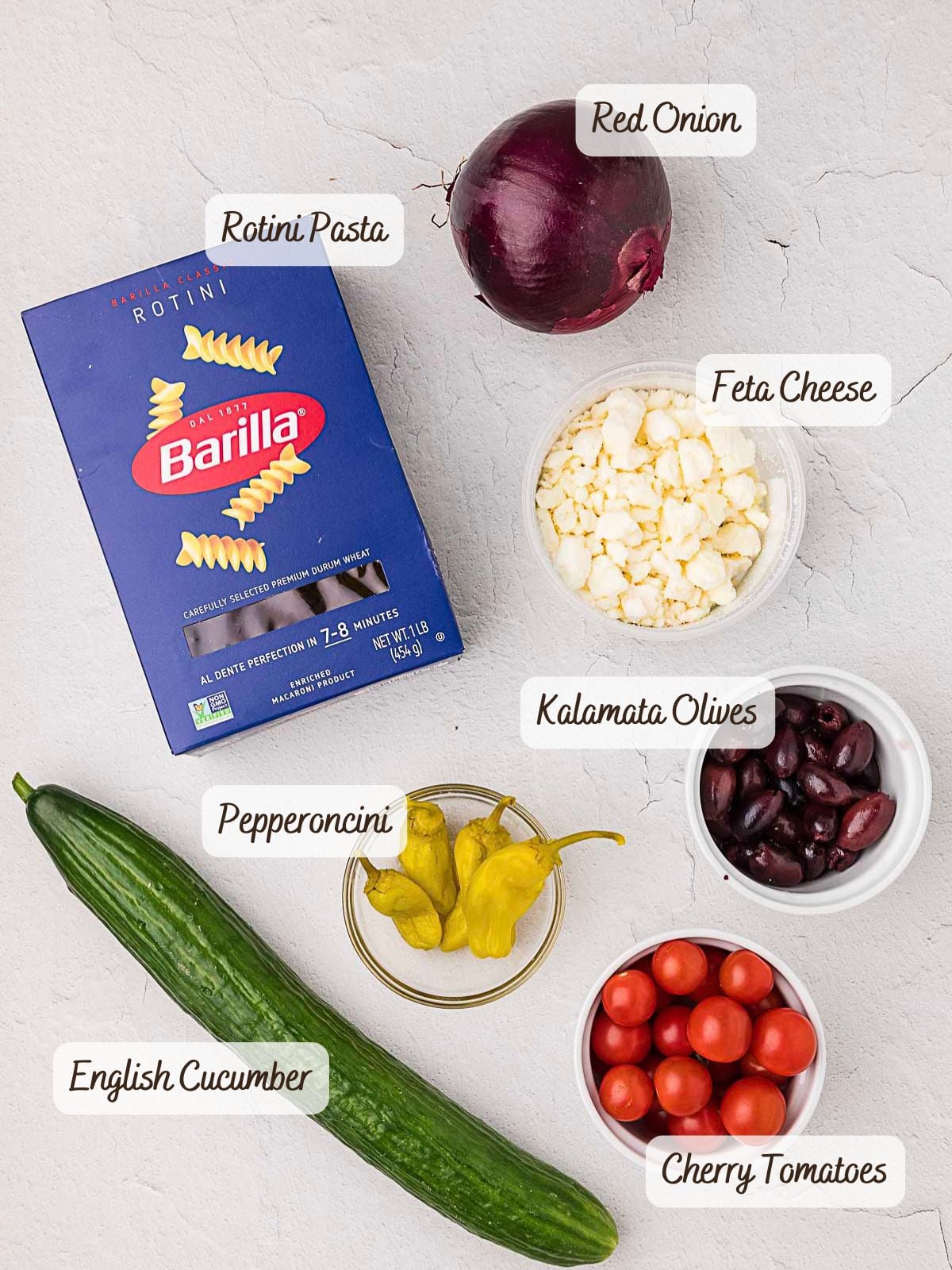 Box of rotini pasta, a whole red onion, feta cheese, kalamata olives, pepperoncini peppers, English cucumber, and cherry tomatoes-everything you need to make a fresh and delicious Greek Pasta Salad.