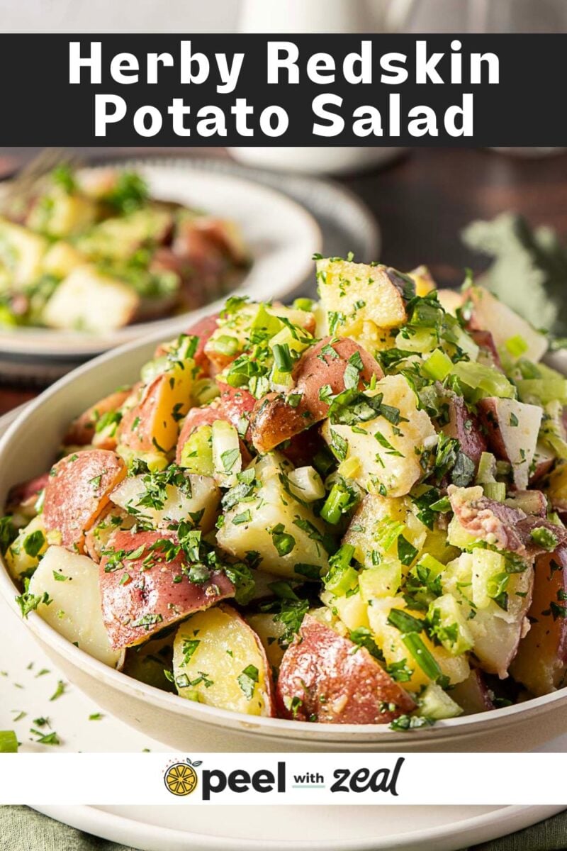 A bowl of herby redskin potato salad garnished with chopped herbs, with another plate of the same salad in the background. Text at the top reads "Herby Redskin Potato Salad.