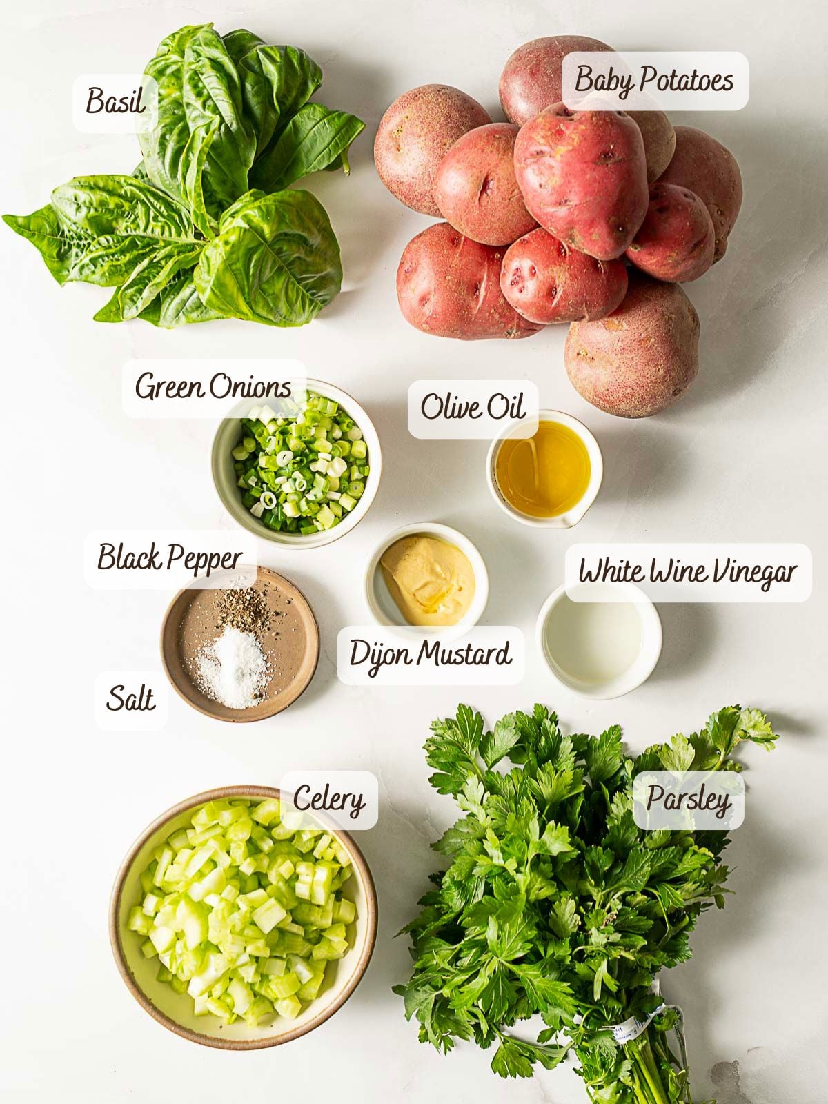 Ingredients for a potato salad recipe displayed on a white surface, including labeled basil, baby potatoes, green onions, olive oil, white wine vinegar, black pepper, salt, Dijon mustard, celery, and parsley.