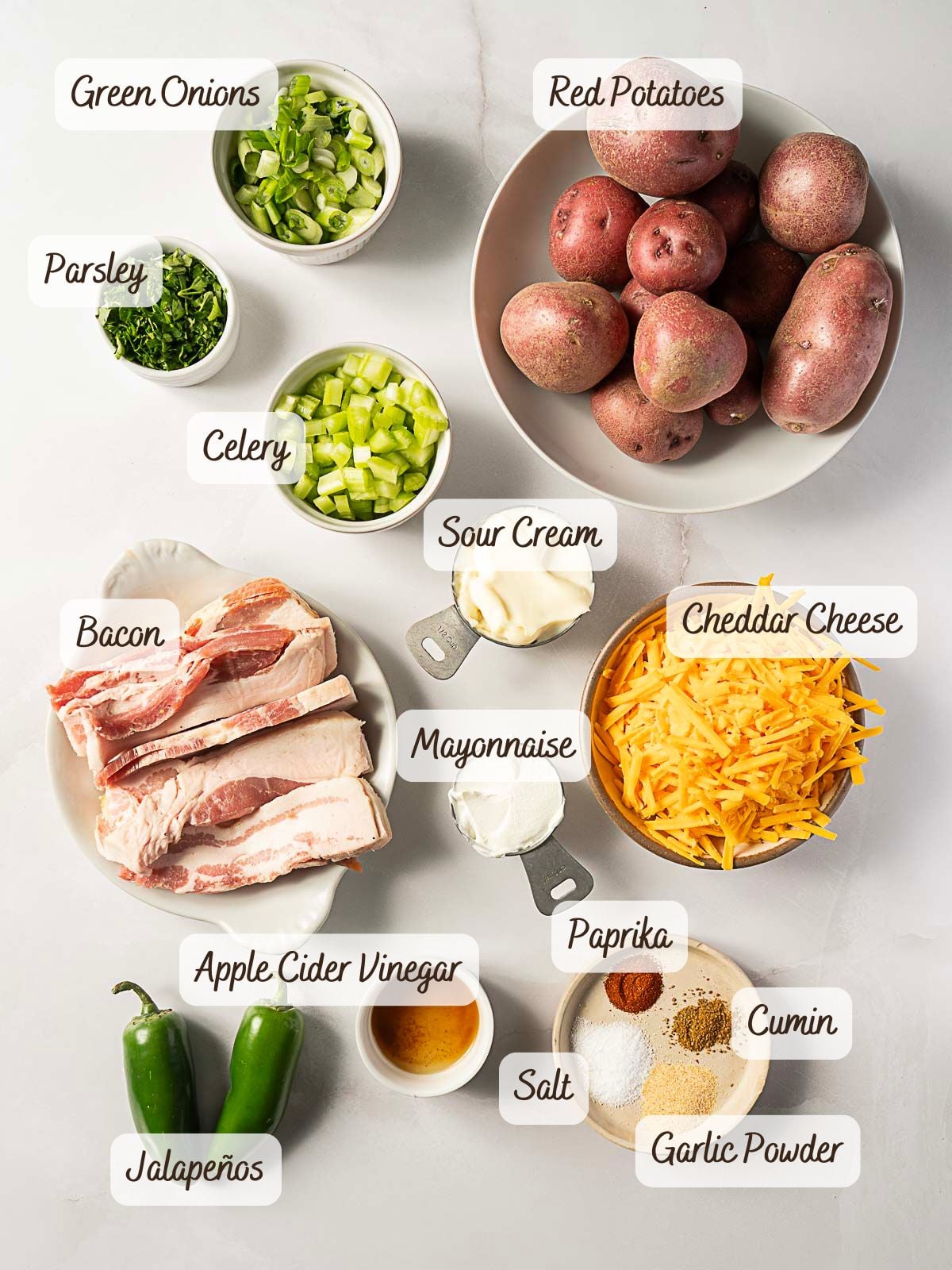 Labeled ingredients for a jalapeno popper potato salad recipe, including red potatoes, green onions, parsley, celery, bacon, sour cream, mayonnaise, cheddar cheese, apple cider vinegar, spices, and jalape&ntilde;os.