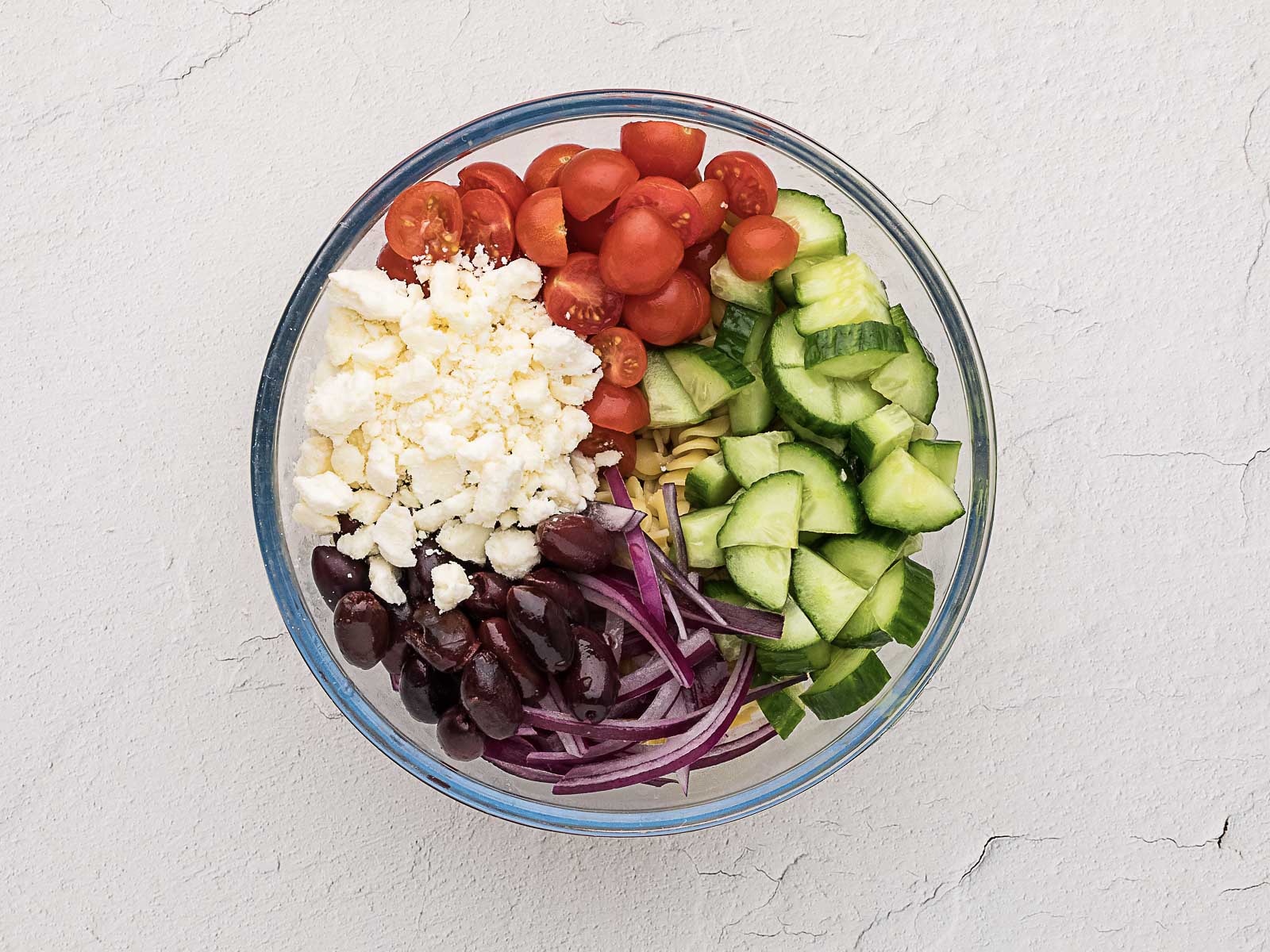 A glass bowl filled with cherry tomatoes, cucumber slices, feta cheese, kalamata olives, red onion, and pasta, arranged in separate sections.