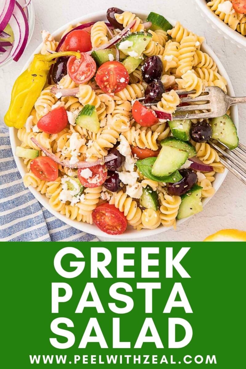 A bowl of Greek pasta salad with rotini, cherry tomatoes, cucumbers, olives, red onion, feta cheese, and a pepper, next to a fork and a blue striped napkin.