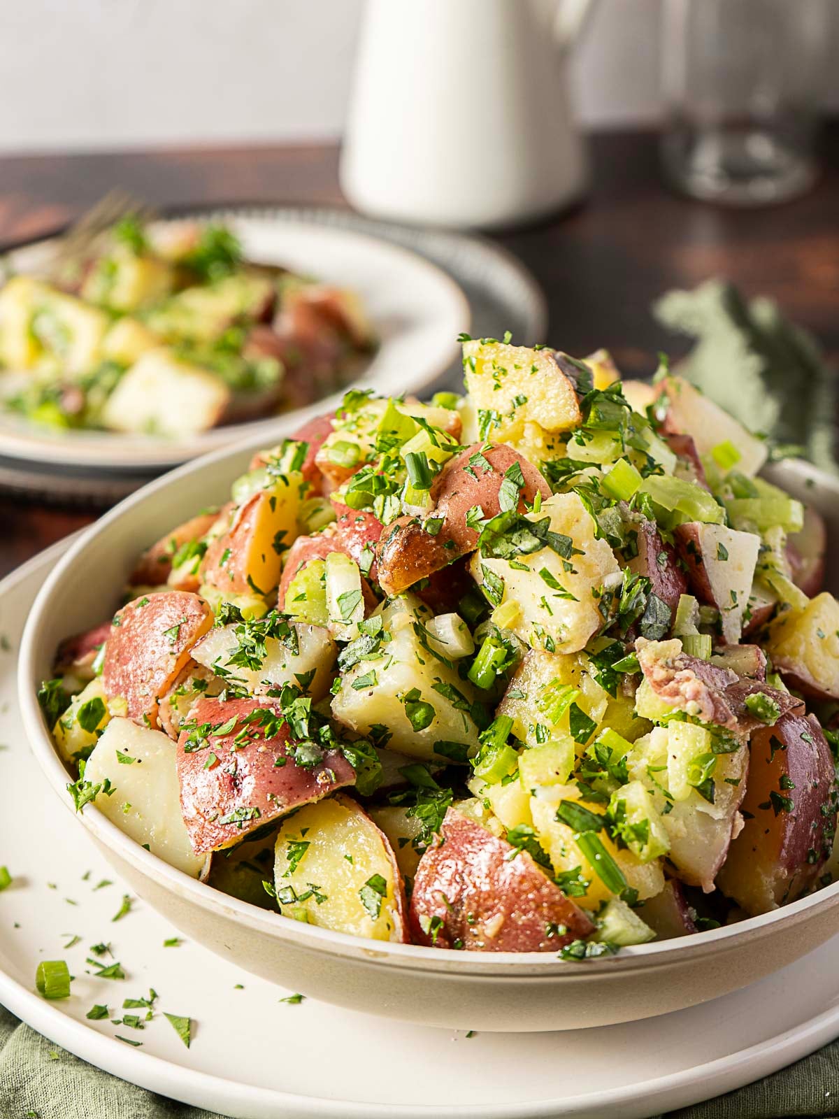 A bowl of potato salad with chopped red potatoes, celery, green onions, and parsley, garnished with fresh herbs. A plate with more salad is in the background.