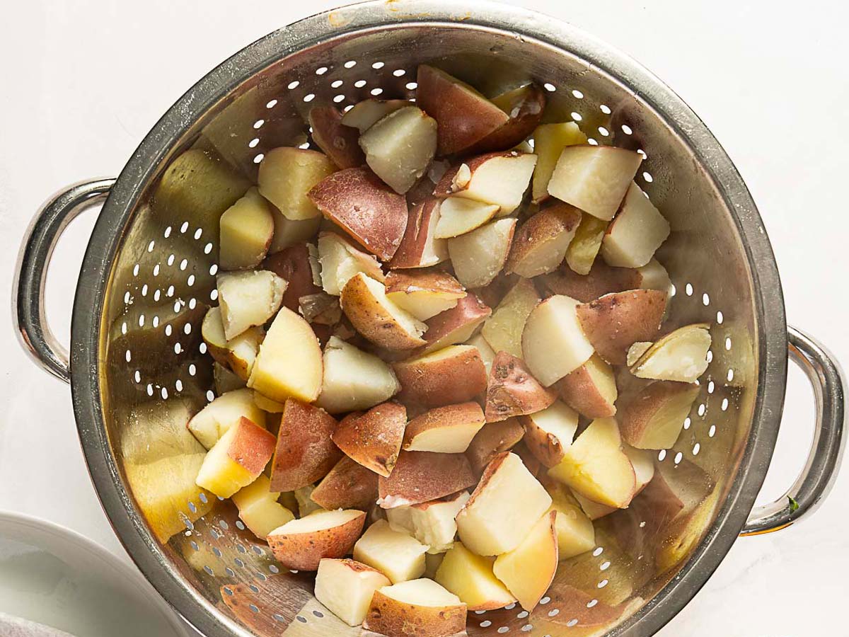 Steamed chopped red potatoes are draining in a metal colander, ready to be tossed into a fresh herbed potato salad.