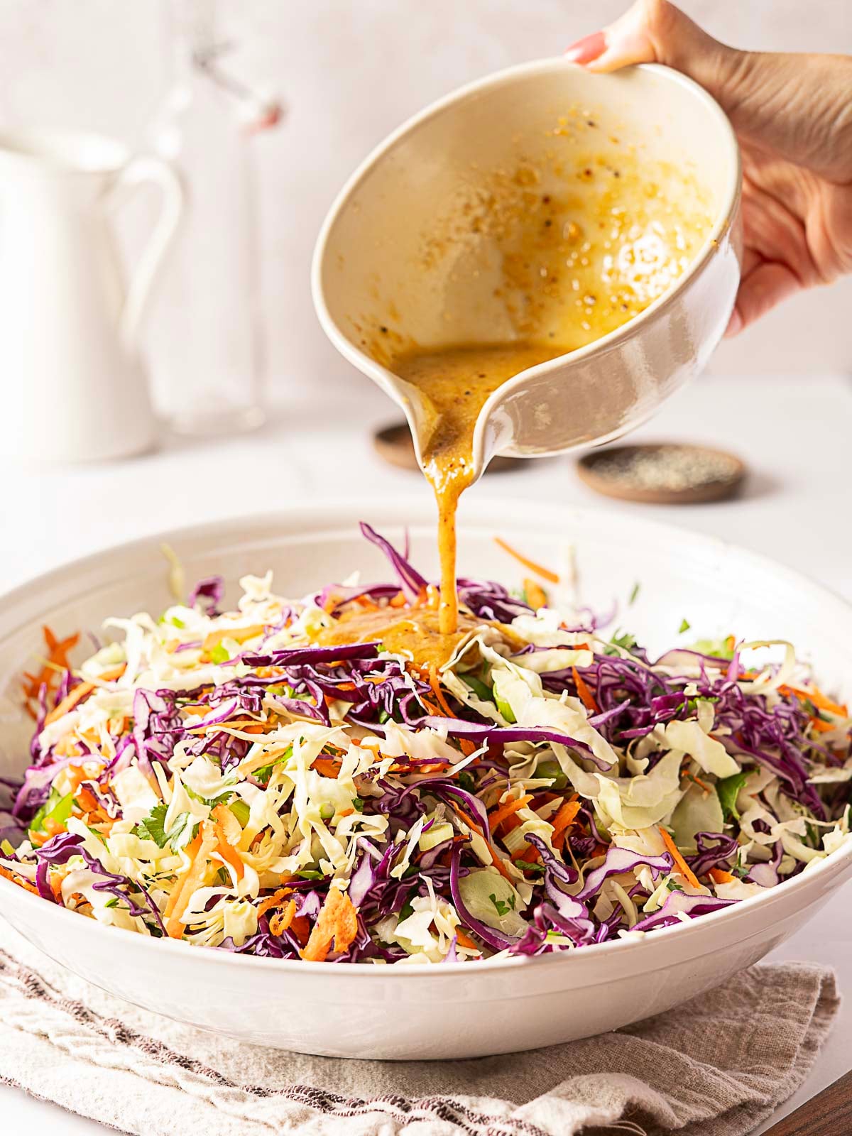 A hand pours dressing from a bowl onto a large salad made of shredded green and purple cabbage, carrots, and herbs, in a white serving dish.