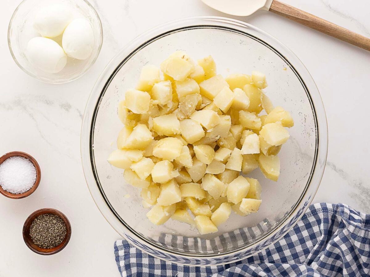 A glass bowl filled with chopped boiled potatoes sits on a counter next to a bowl of hard-boiled eggs, small bowls of salt and pepper, a spatula, and a blue checkered cloth.