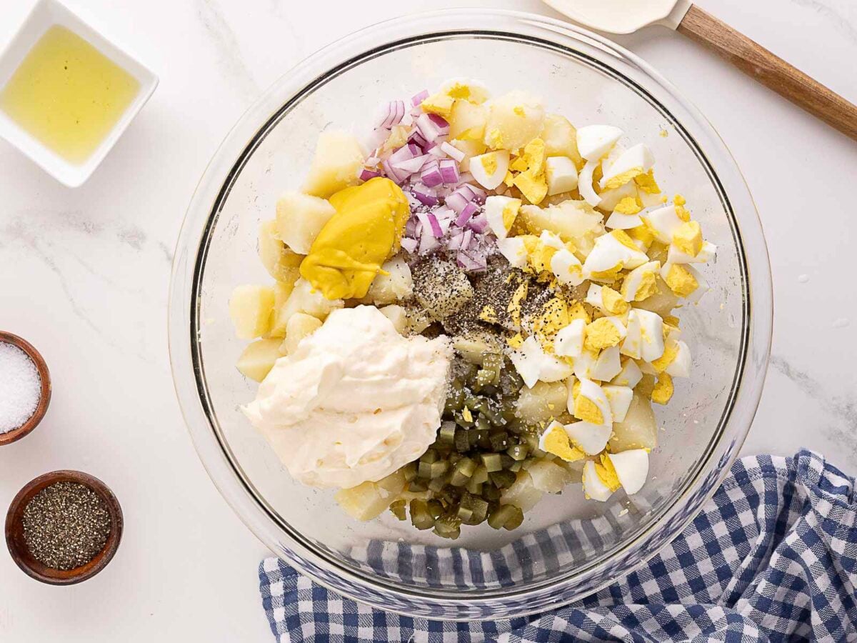A glass bowl with chopped potatoes, hard-boiled eggs, red onions, mayonnaise, mustard, chopped pickles, salt, and pepper on a countertop with a blue checkered napkin.