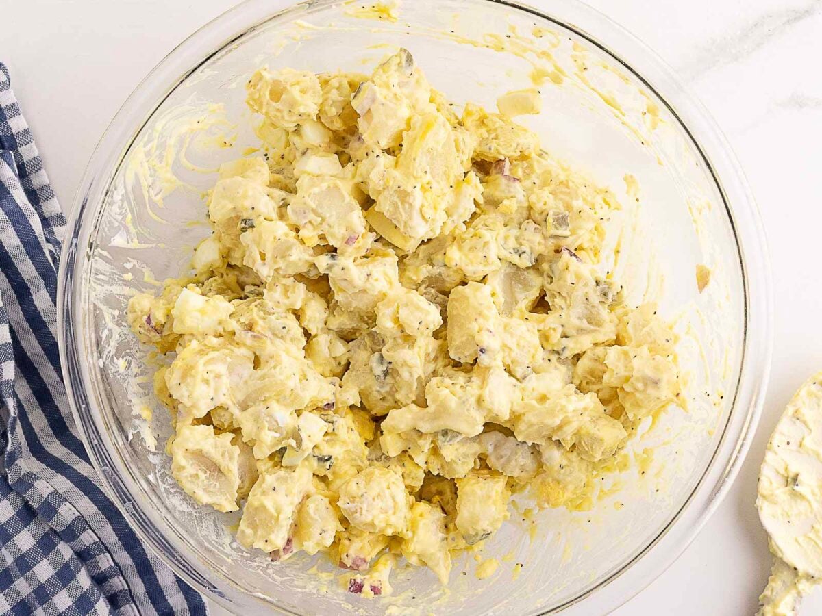 A glass bowl filled with potato salad mixed with mayonnaise and seasonings sits on a white surface next to a blue-and-white striped cloth.