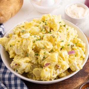 A bowl of potato salad with chopped chives and red onions on top, placed on a wooden board next to a blue plaid napkin.