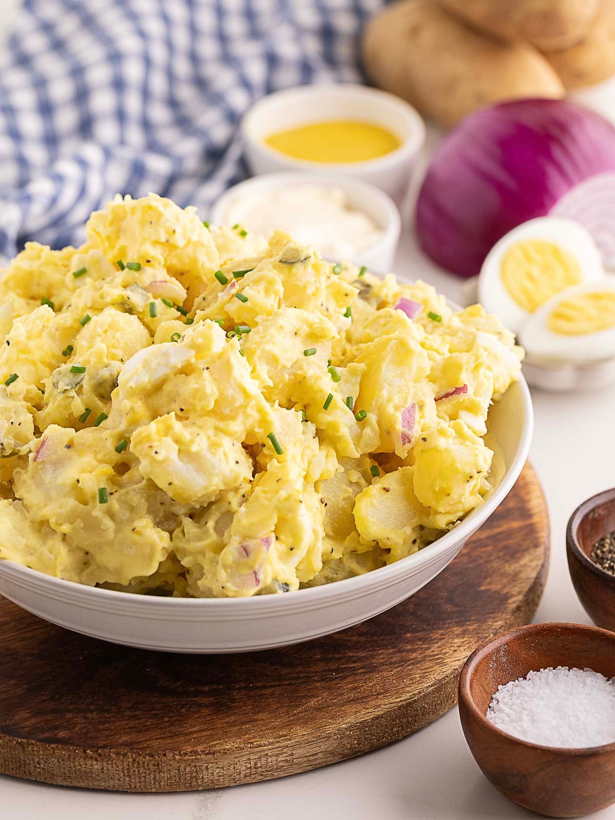 A bowl of potato salad with chopped chives, red onion, and hard-boiled eggs, surrounded by condiments, a red onion, potatoes, and a blue checkered cloth.