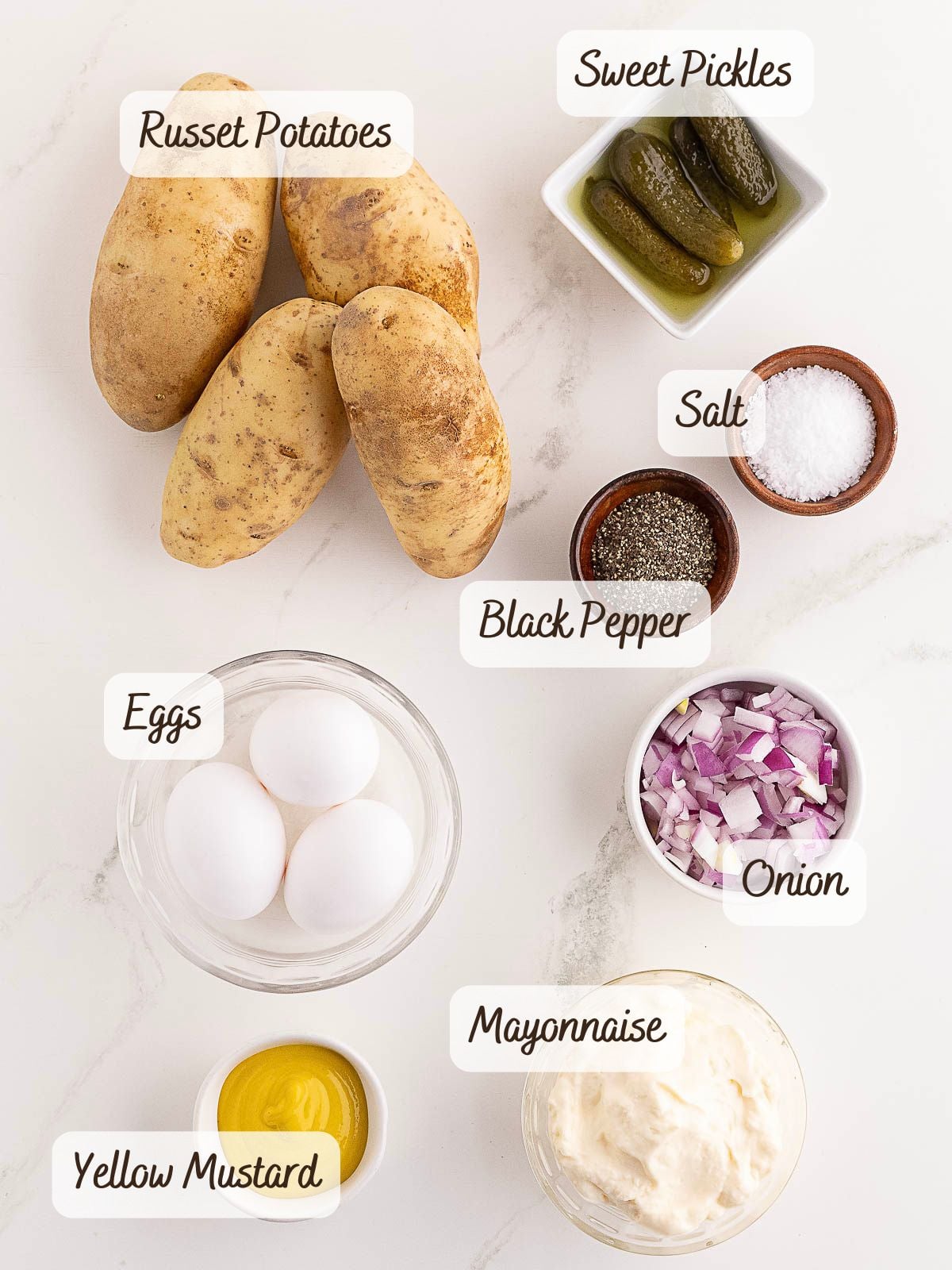 Ingredients for Southern Potato Salad laid out on a white surface: russet potatoes, sweet pickles, salt, black pepper, eggs, chopped onion, yellow mustard, and mayonnaise.