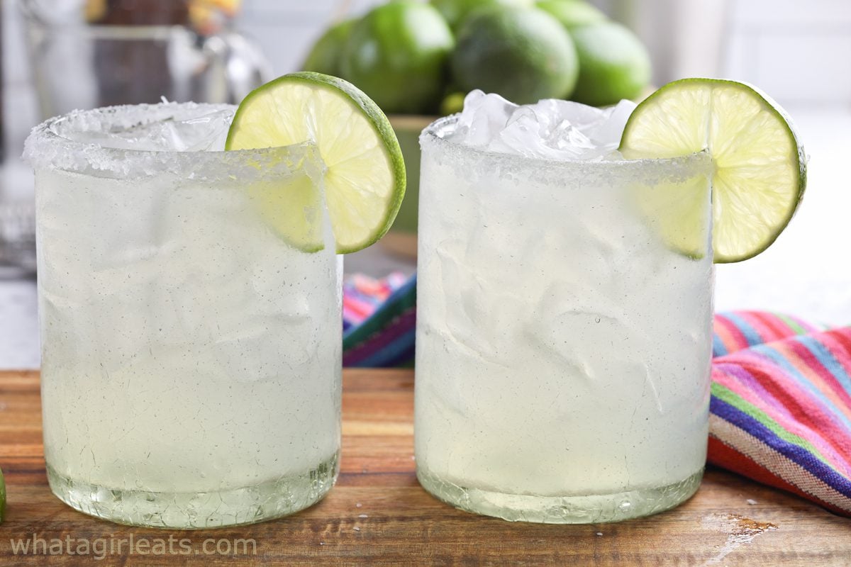 Two salt-rimmed glasses filled with ice and a clear drink, each garnished with a lime slice, sit on a wooden surface with limes and a colorful cloth in the background.