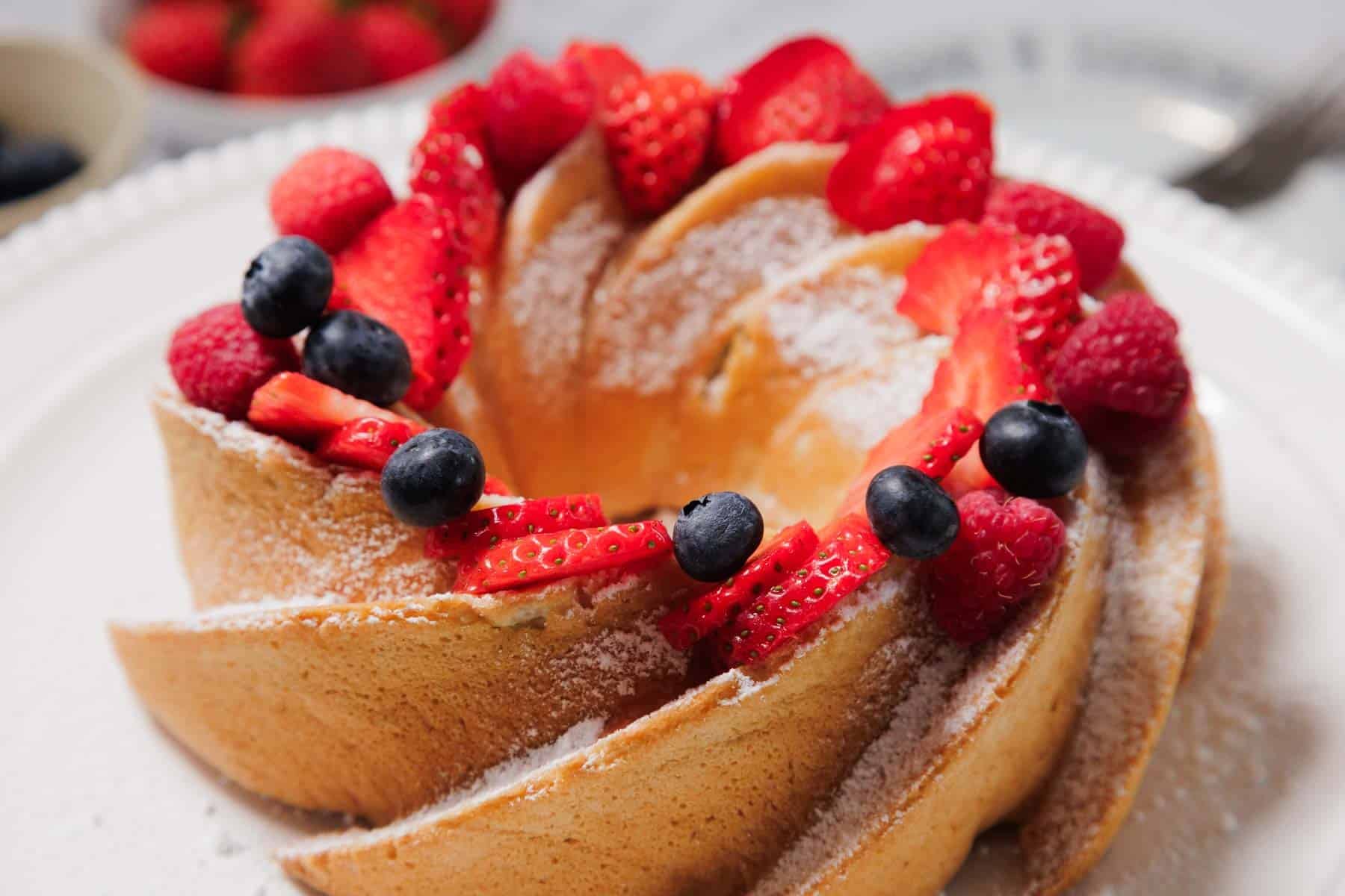 A Bundt cake dusted with powdered sugar, decorated with sliced strawberries, blueberries, and raspberries, displayed on a white plate.