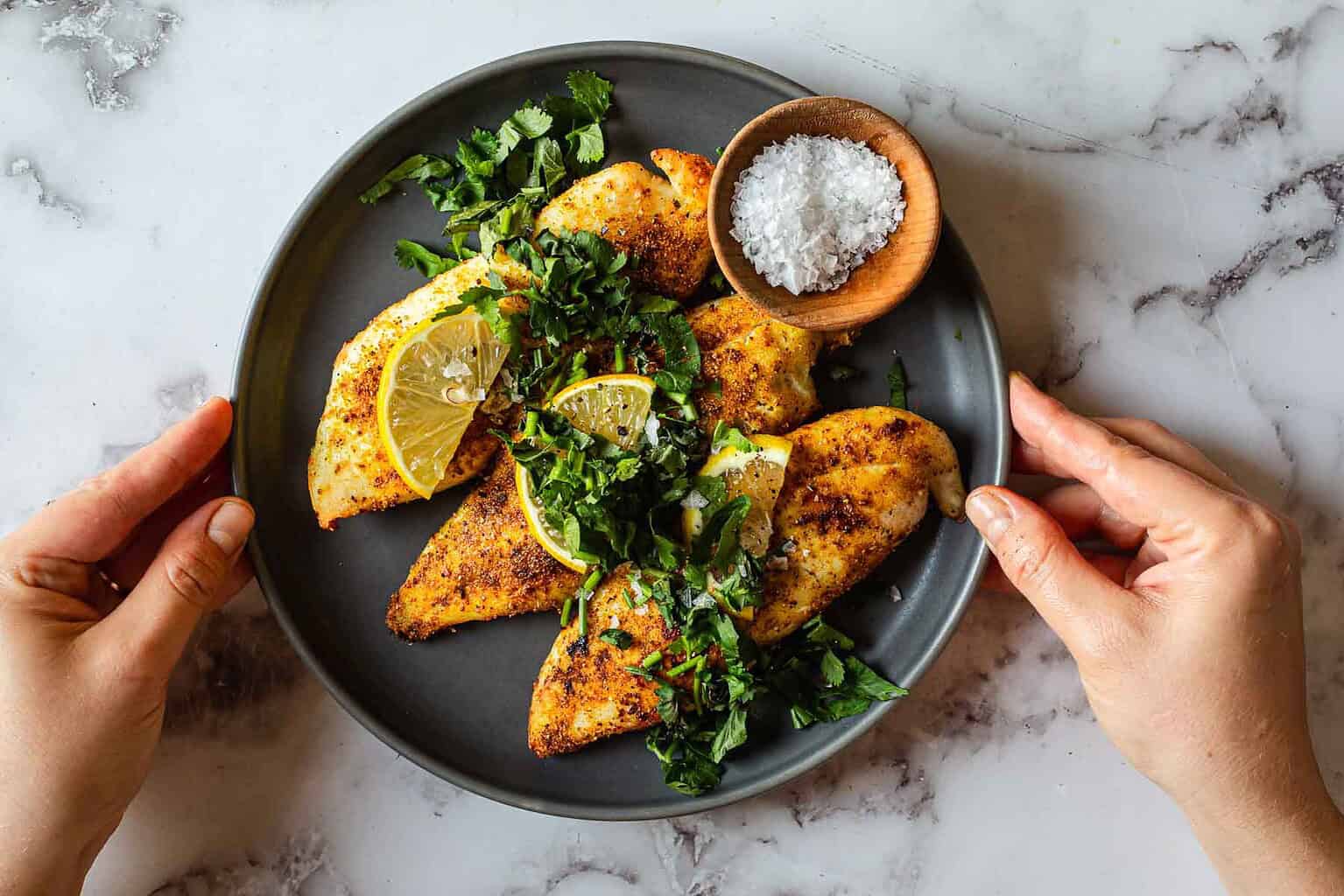 A plate of seasoned grilled chicken breasts garnished with lemon slices and fresh herbs, with a small bowl of sea salt on the side. Two hands hold the plate on a marble surface.