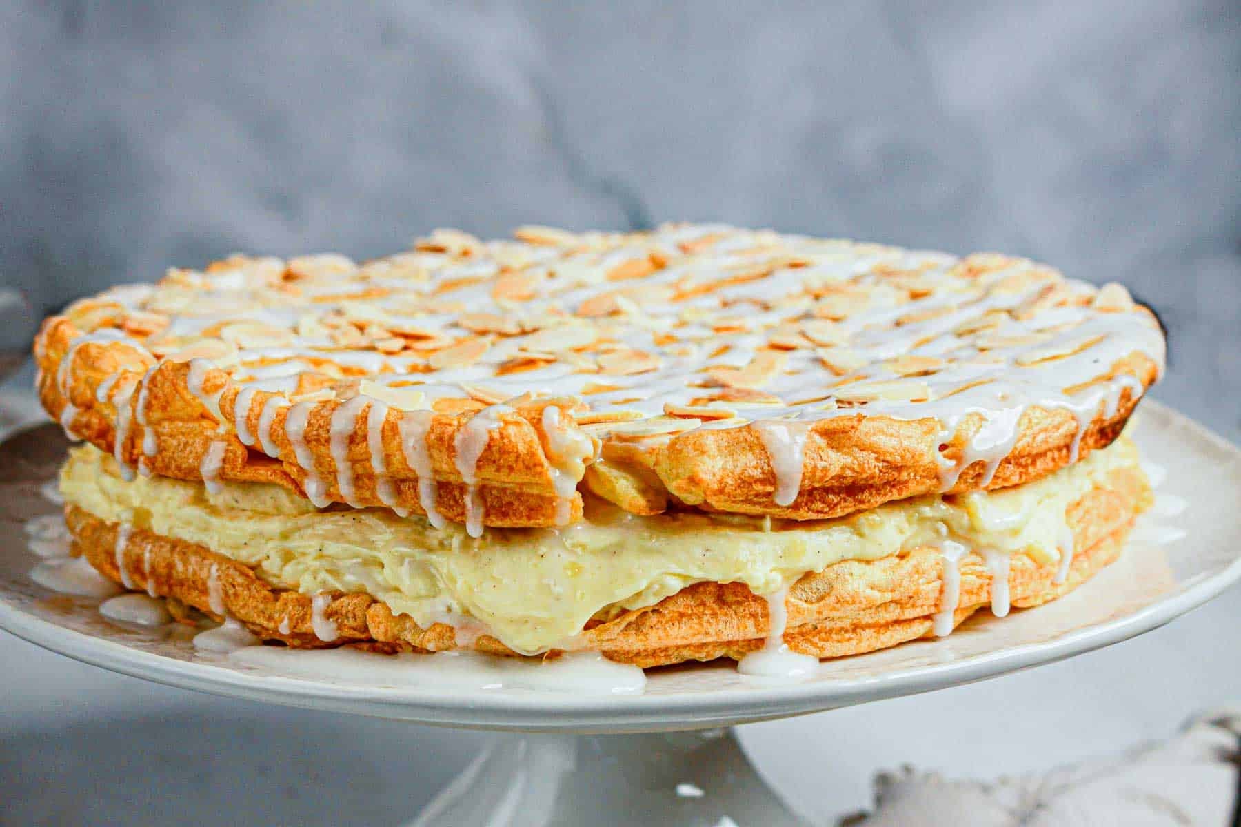 A round layered pastry filled with cream and topped with slivered almonds and white icing, displayed on a white cake stand.