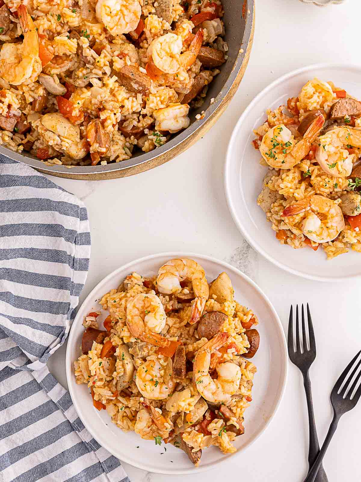 Plates of shrimp jambalaya with rice, sausage, chicken, and vegetables are served next to a pan and two black forks on a white surface.