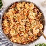 A skillet filled with shrimp, sausage, chicken, rice, and vegetables, stirred with a wooden spoon.