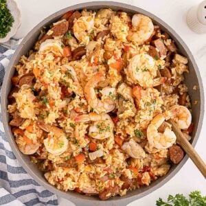 A skillet filled with shrimp, sausage, chicken, rice, and vegetables, stirred with a wooden spoon.