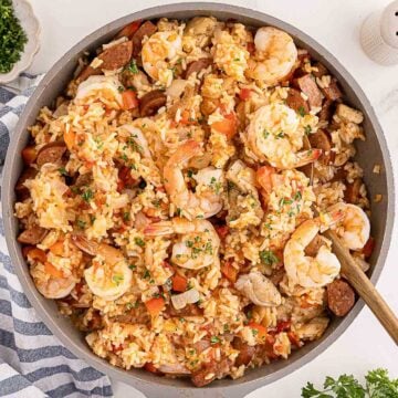 A skillet filled with shrimp, sausage, chicken, rice, and vegetables, stirred with a wooden spoon.