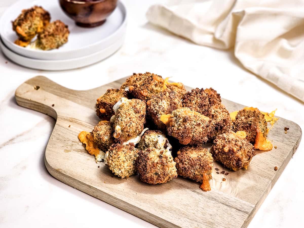 A pile of breaded, baked broccoli and cheese bites on a wooden cutting board, with a plate and a bowl in the background.