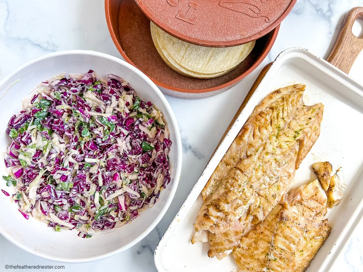 A bowl of cabbage slaw, a stack of corn tortillas in a tortilla warmer, and a tray with cooked fish fillets on a white surface.