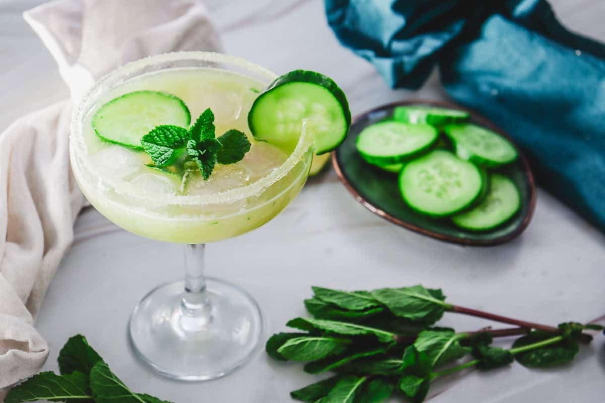 A glass of cucumber cocktail garnished with mint and cucumber slices, with fresh mint leaves and a plate of cucumber slices nearby on a white surface.