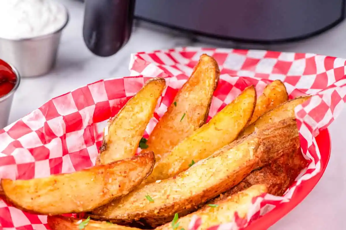 Basket of seasoned potato wedges on red checkered paper, served with dipping sauces in metal cups in the background.