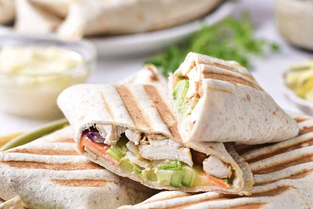 Grilled chicken wraps cut in half, showing lettuce, cucumber, and other vegetables inside, stacked on a plate with a bowl of sauce in the background.