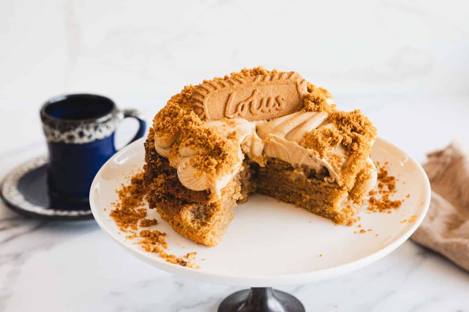 A round cake topped with Lotus Biscoff cookies and crumbs, sitting on a white cake stand, with a dark cup and saucer in the background.