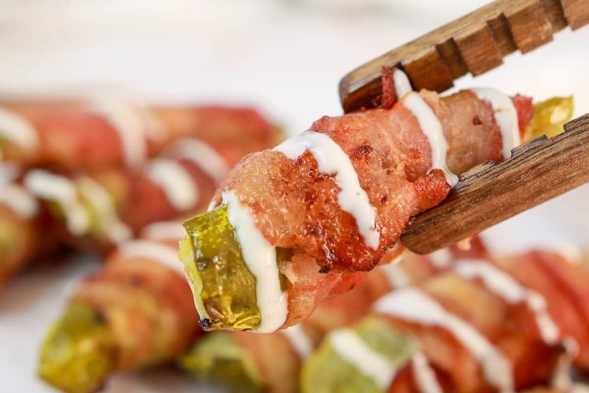 Close-up of chopsticks holding a bacon-wrapped pickle spear drizzled with a white sauce, with more bacon-wrapped pickles in the blurred background.