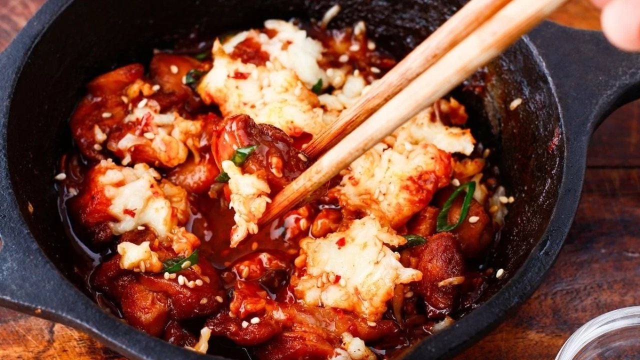 A cast iron pan filled with spicy, saucy chicken pieces topped with melted cheese, garnished with sesame seeds and chopped green onions; chopsticks are picking up a piece.