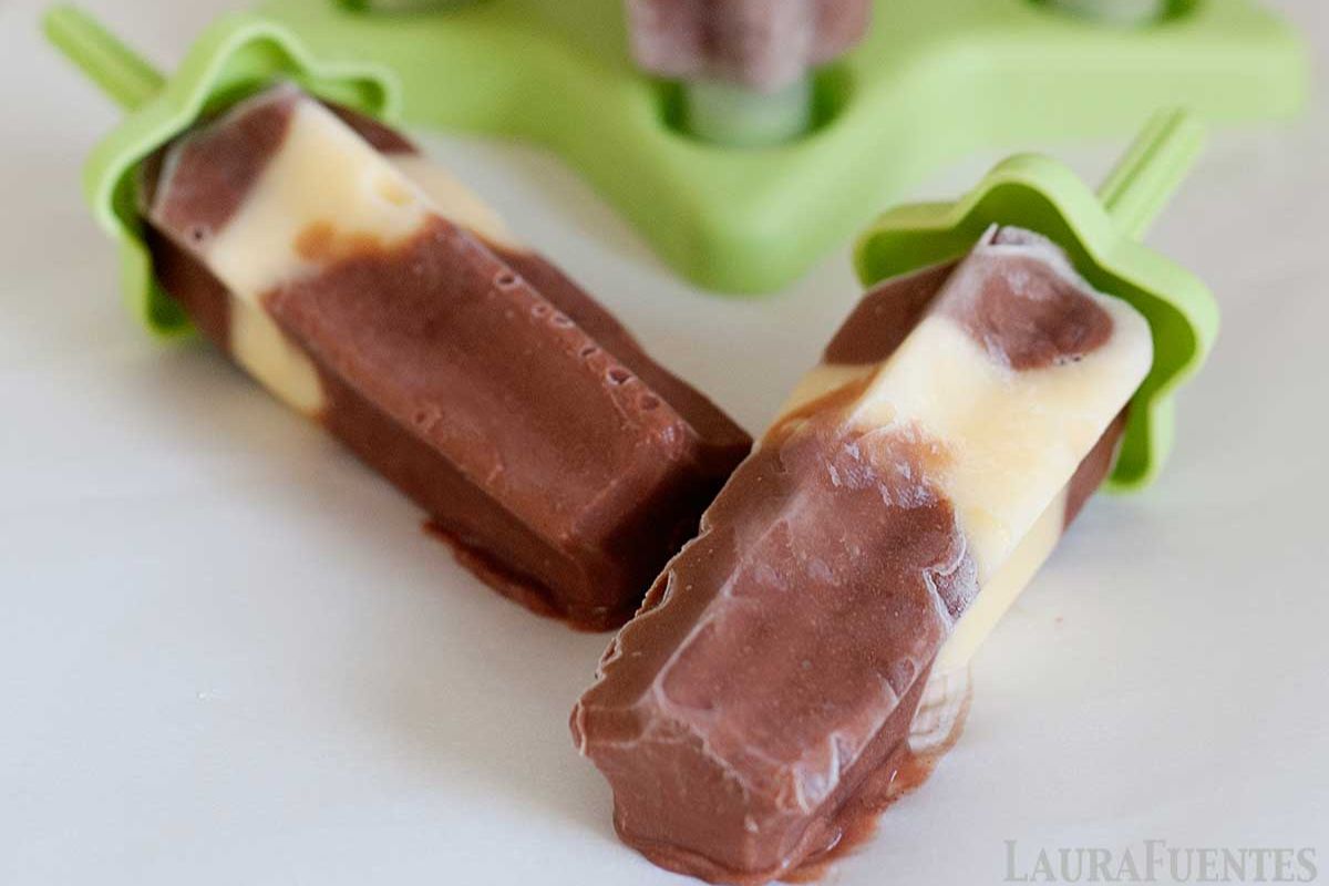 Two chocolate and vanilla layered popsicles with green handles are melting on a white surface. A popsicle mold is visible in the background.