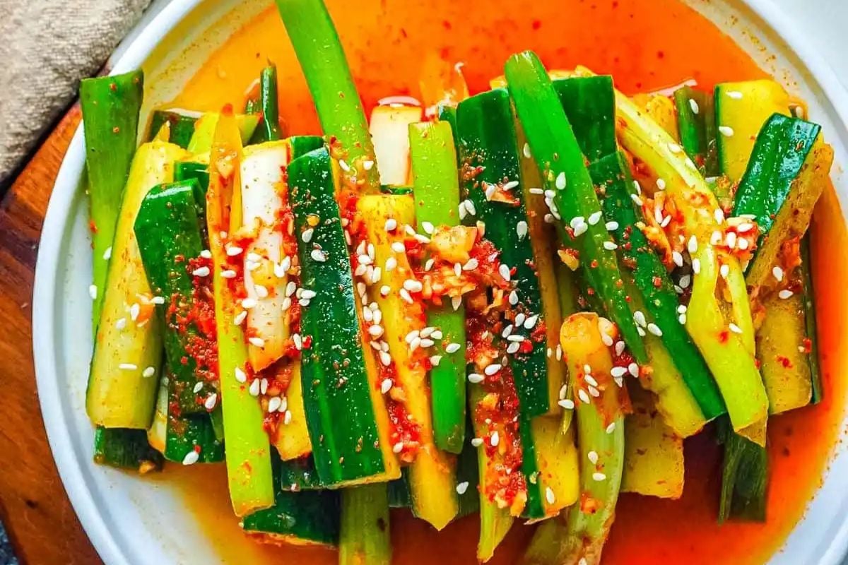 Sliced cucumbers seasoned with red chili, sesame seeds, and spices, arranged on a white plate with a red-orange sauce.