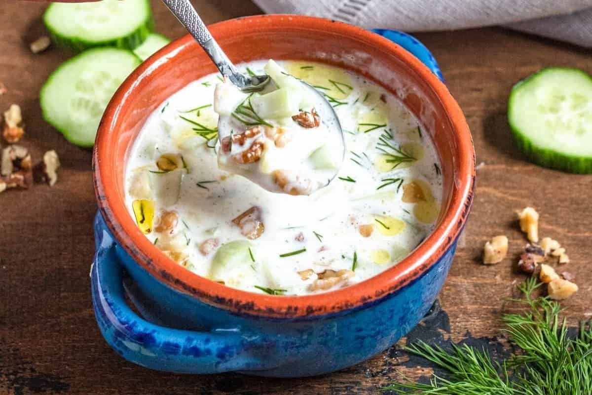 A blue bowl of creamy cucumber soup garnished with fresh dill and walnuts, with a spoon lifting a portion. Sliced cucumbers and walnuts are scattered on a wooden surface nearby.
