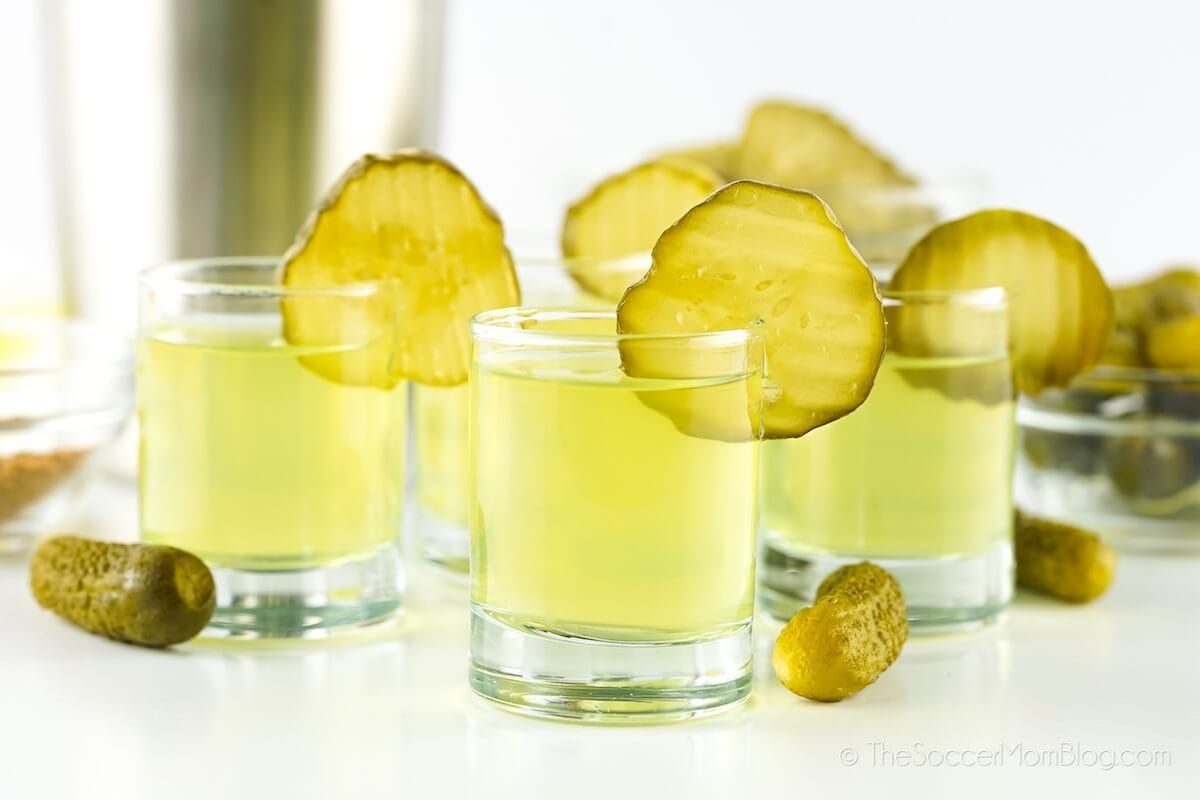 Four shot glasses filled with a yellow-green liquid, each garnished with a pickle slice, with small pickles and blurred glassware in the background.