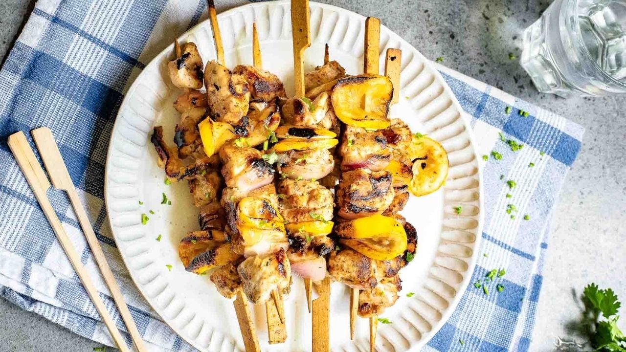 Grilled chicken and vegetable skewers on a white plate, placed on a blue and white checkered cloth with a glass of water nearby.