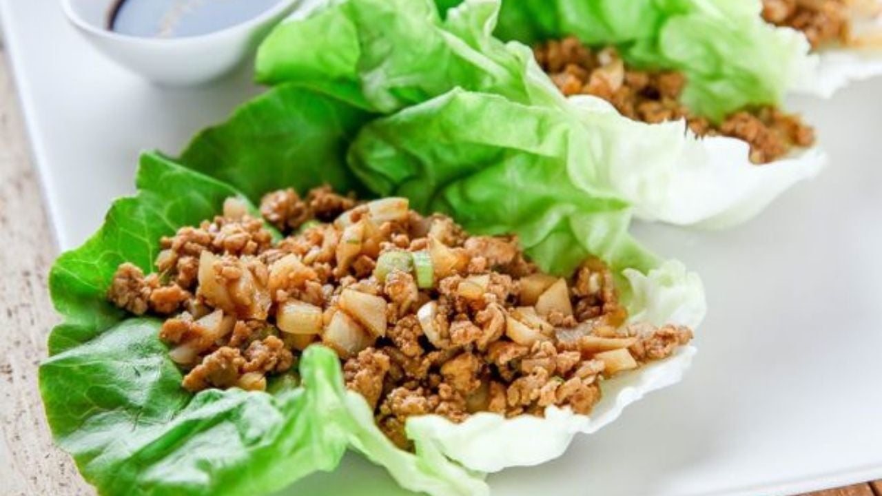 Three lettuce leaves filled with seasoned ground meat and chopped vegetables are arranged on a white plate, with a small bowl of sauce in the background.
