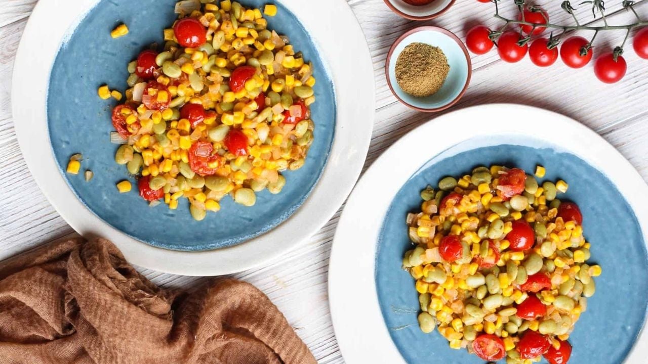 Two blue plates filled with corn, lima beans, and cherry tomatoes, served on a white wooden surface.