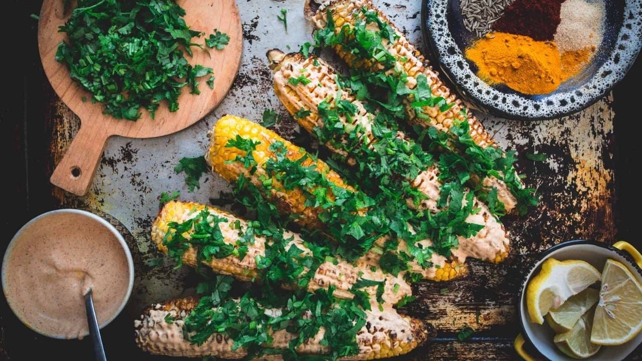 Grilled corn on the cob topped with chopped herbs and a creamy sauce, served with lemon wedges and a spice blend.