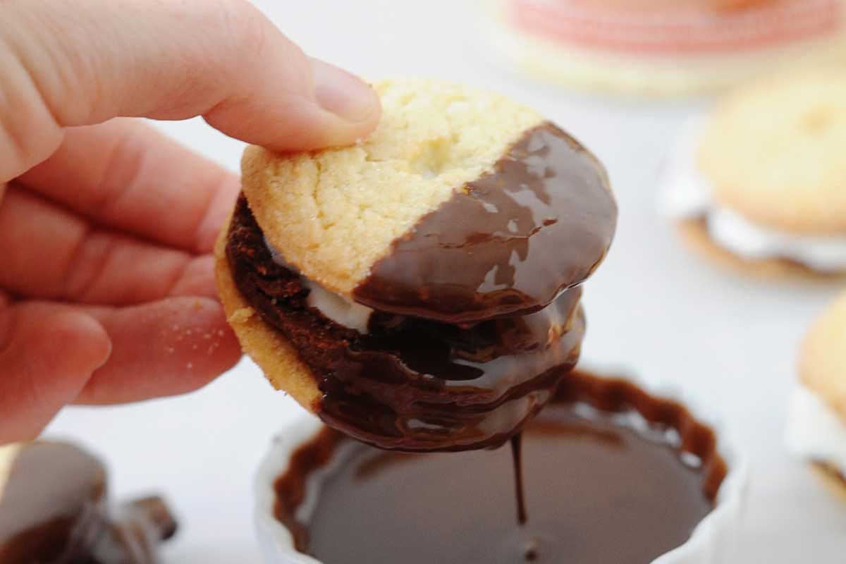 A hand holds a sandwich cookie partially dipped in melted chocolate, with chocolate dripping into a small white bowl below.