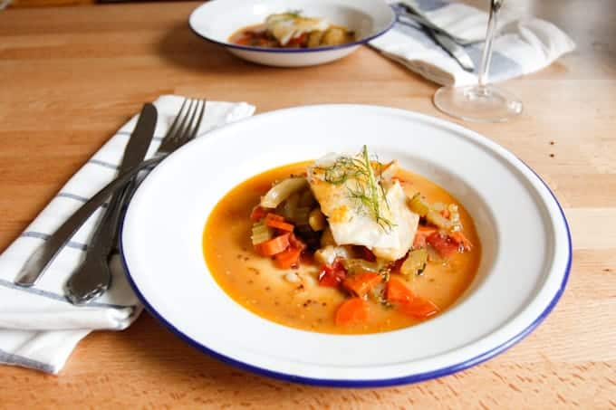 A white plate of fish with vegetables in a broth, with cutlery and another plate of food in the background.