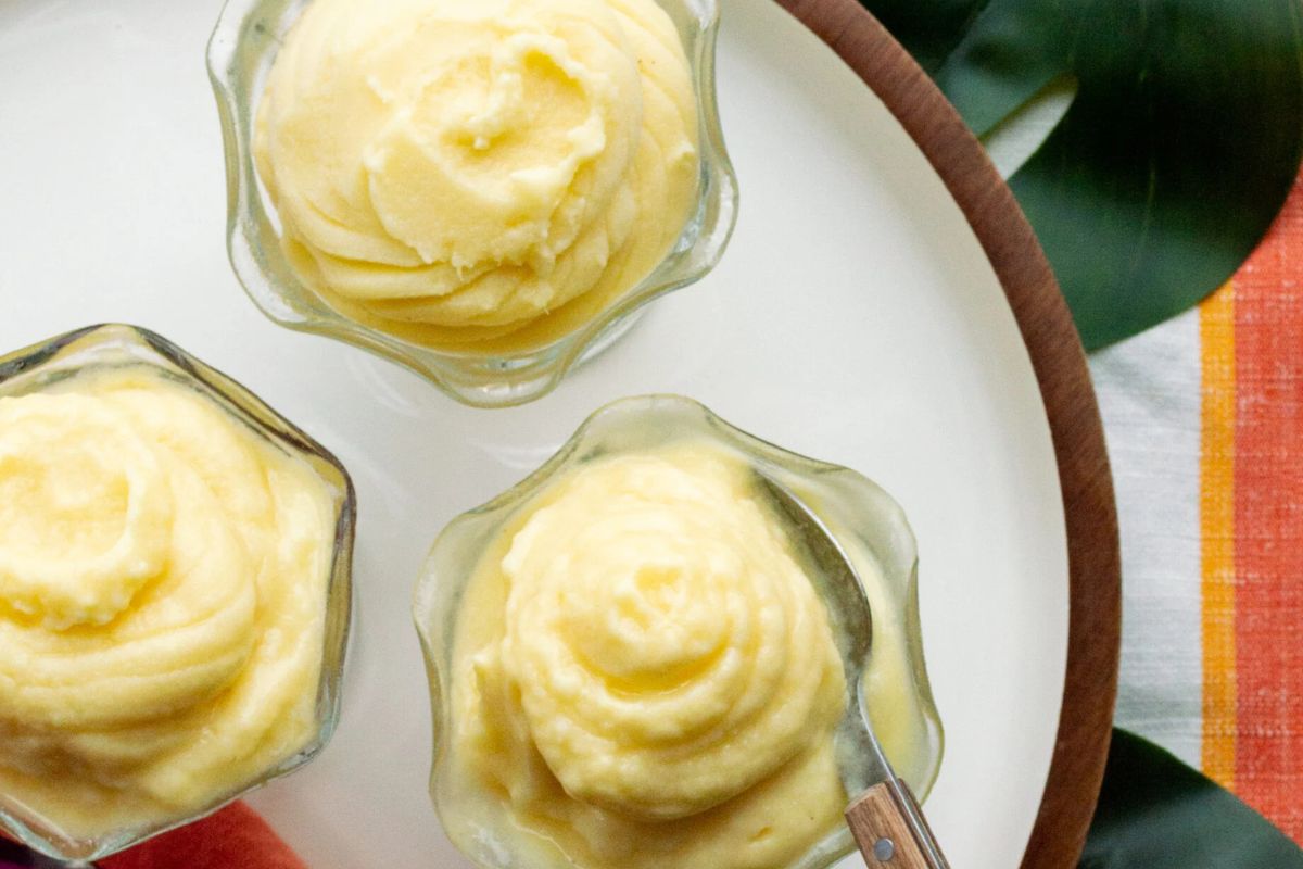 Three glass bowls of creamy yellow dessert, possibly Dole Whip or soft serve, sit on a white plate with a spoon in one bowl. A green leaf and striped cloth are partially visible.