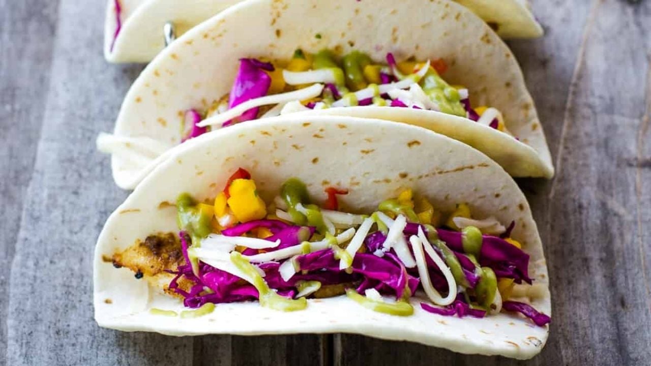 Three tacos filled with fish, red cabbage, corn, and a green sauce.