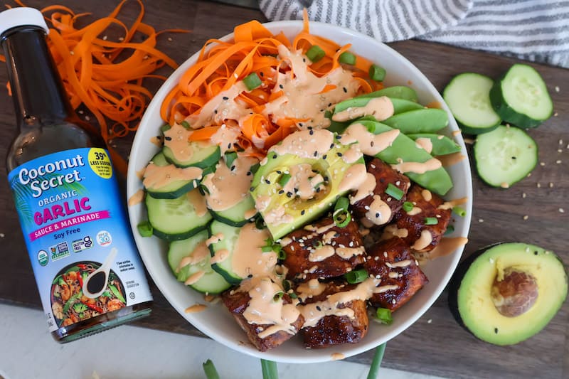 A bowl with sliced avocado, cucumber, carrot ribbons, marinated tofu, and sauce, next to a bottle of Coconut Secret Organic Garlic Sauce & Marinade on a wooden board.