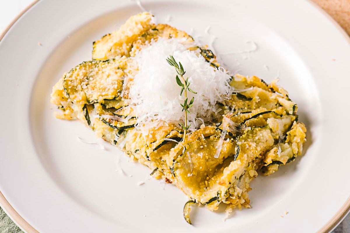 A serving of baked zucchini slices topped with grated cheese and garnished with thyme, this dish brings the fresh flavors of Italian summer recipes to your table on a simple white plate.